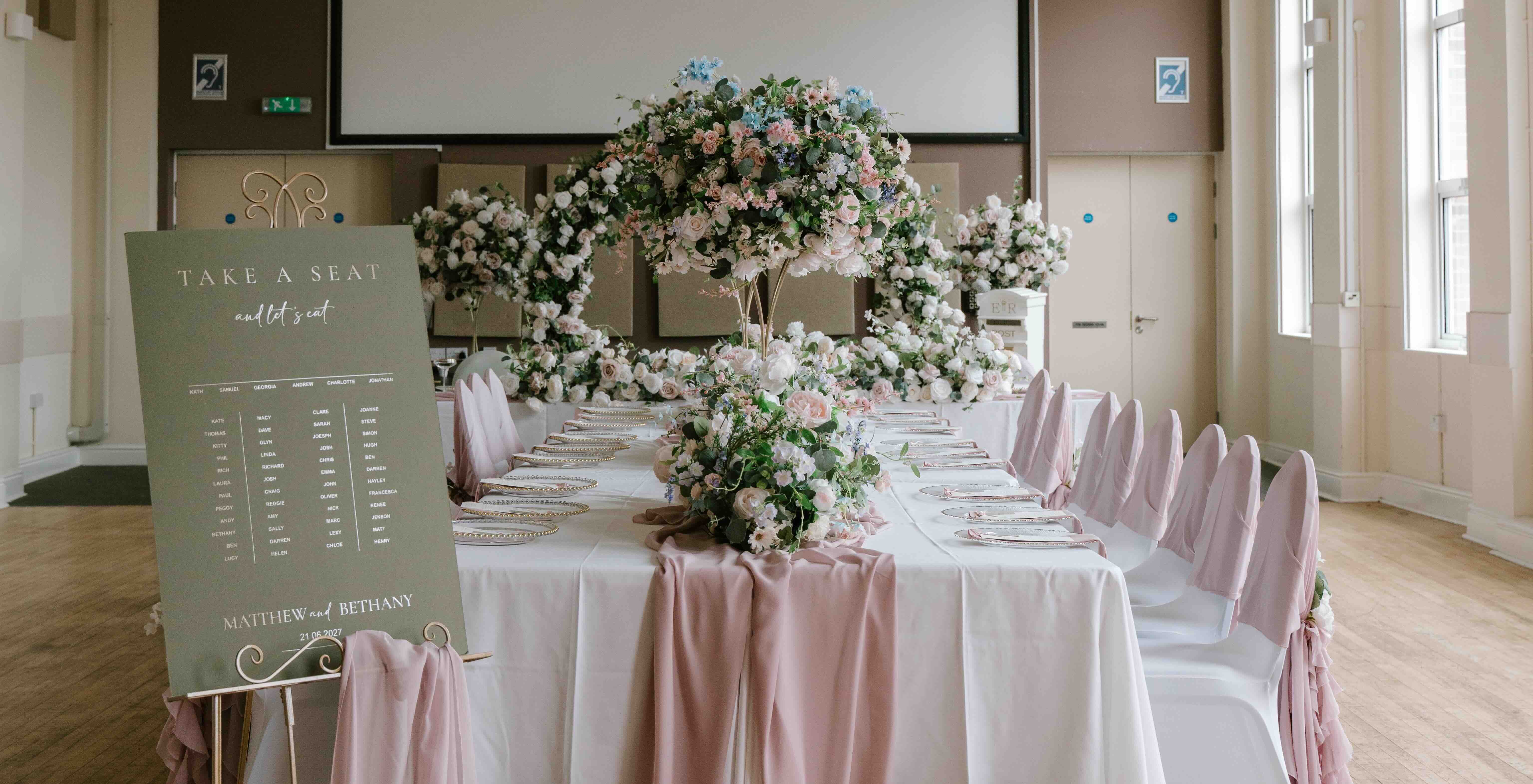 The main hall set for a wedding breakfast, with a seating plan board and tall floral displays