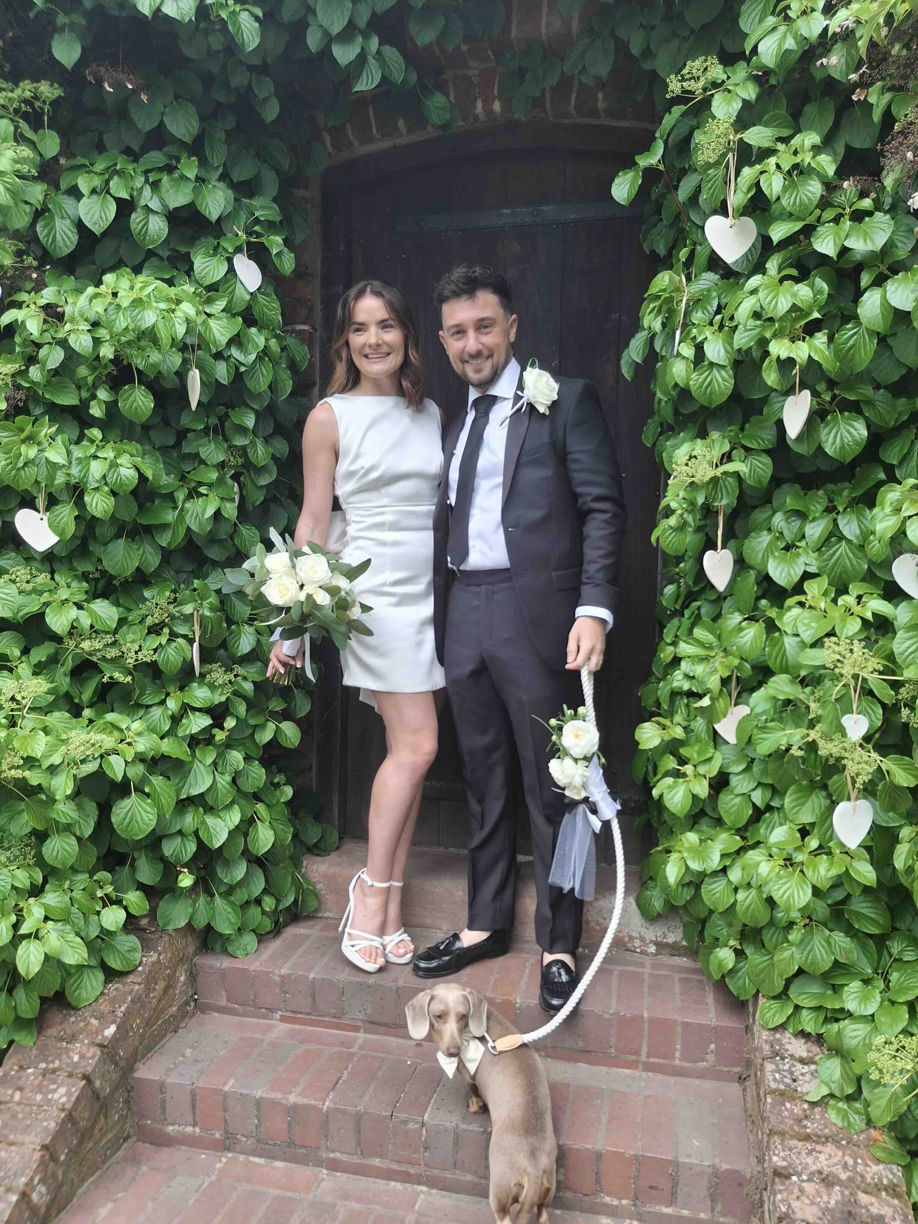 A bride and groom standing in a doorway at Bewdley Museum, surrounded by green foliage and hanging white hearts